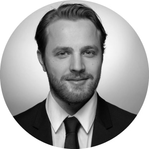 Alt text: A professional portrait of a bearded man with slicked, parted hair; he smiles gently while wearing a smart suit and neatly tied tie. A plain black and white background puts emphasis on his face, symbolizing the professionalism in Jonas Construction Software.