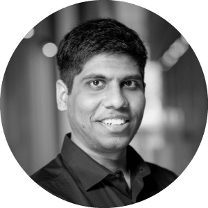 Alt text: A smiling South Asian man with black hair, dressed in a black Jonas Construction shirt, looking directly at the camera against an indoor blurred background. His friendly demeanor reflects the welcoming culture of Jonas Construction Software.