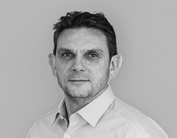 Black-and-white portrait of a man with short, dark hair, wearing a light-colored button-up shirt. He is part of the leadership team and looks directly at the camera with a neutral expression. The background is plain light gray.