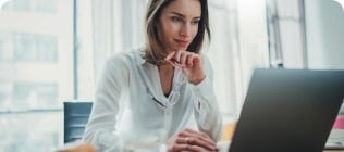 Alt text: A woman with long brown hair is seated at a table, focused on a laptop screen. She is holding her glasses in one hand and is dressed in a white blouse. The setting is a bright room featuring large windows in the background, with papers and a drink placed on the table.