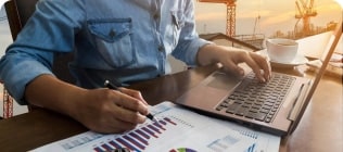 Alt text: A person wearing a denim shirt sits at a desk, working on a laptop while holding a pen. They are examining graphs and charts on a sheet of paper. A cup of coffee is placed nearby. In the background, cranes are silhouetted against a sunset, indicating an industrial or construction site setting.